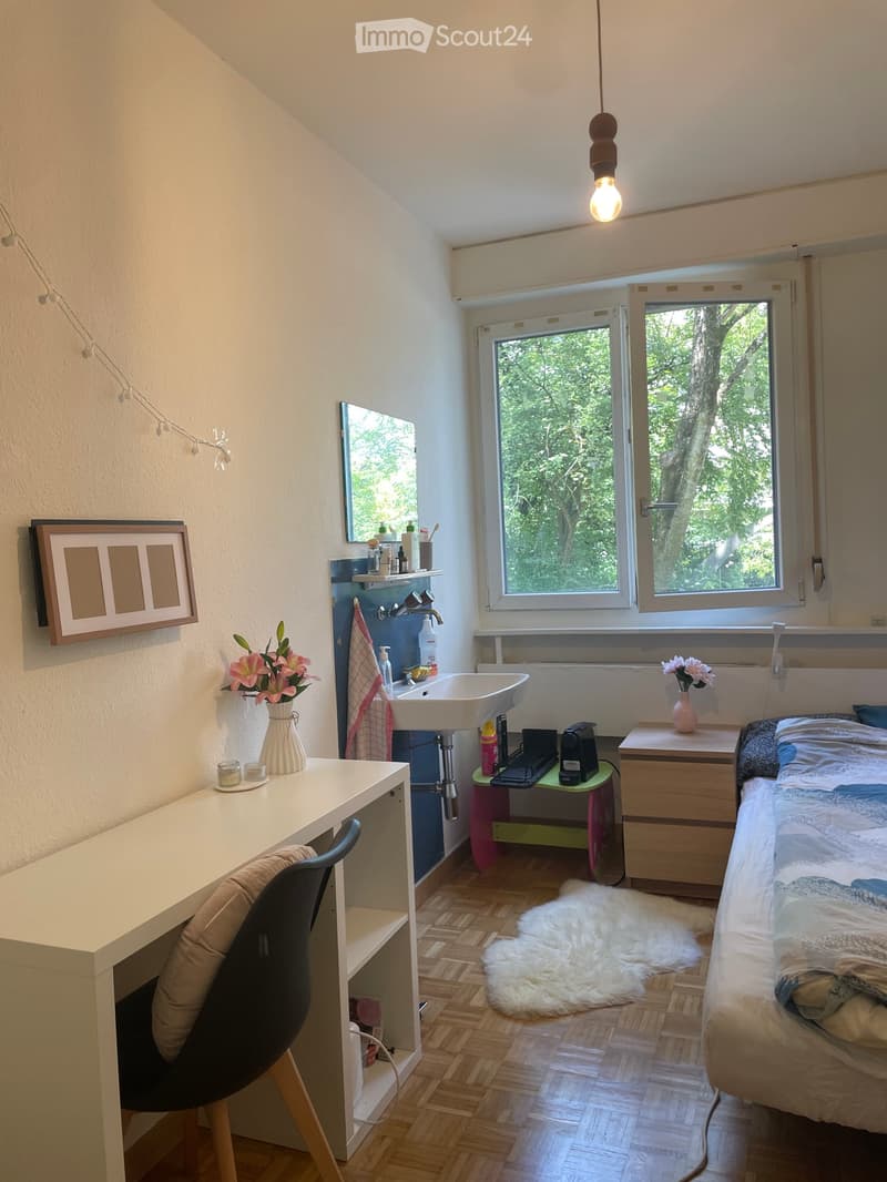 Cozy bedroom with a bed, desk, chair, sink, mirror, framed picture, vase with flowers, furry rug, nightstand, and a small table with a cup and other items.