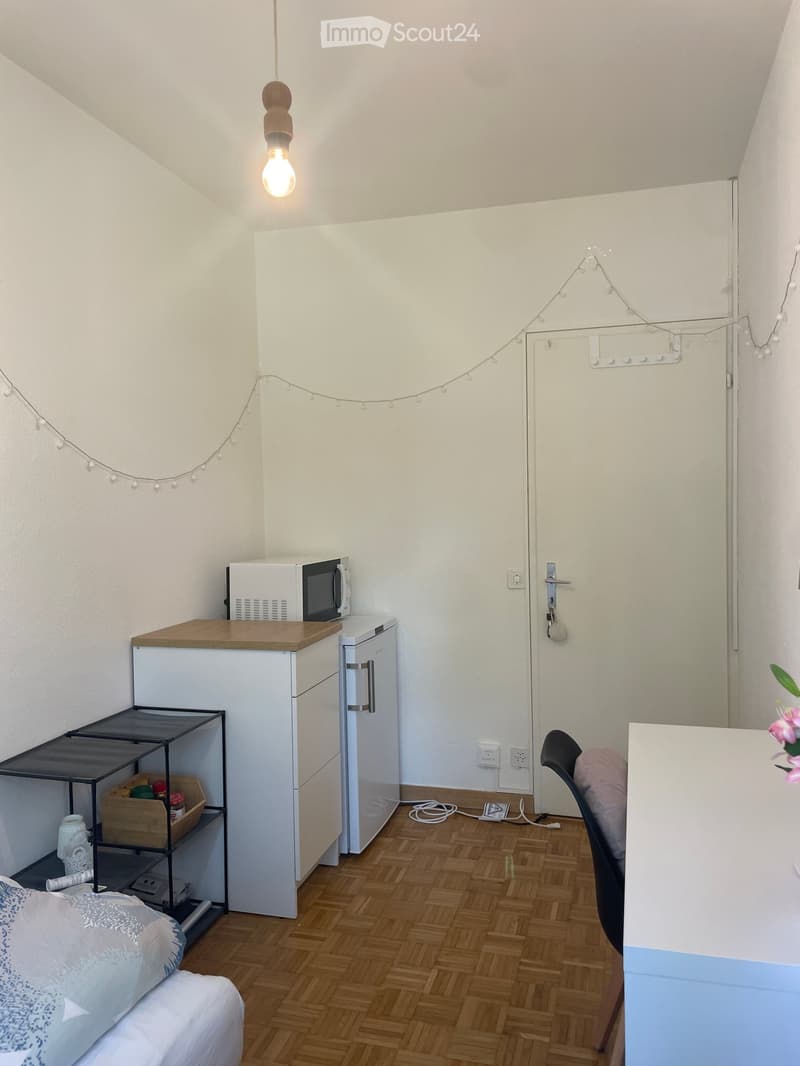 A small, furnished room with a bed, a refrigerator, a microwave, a chair, a small table, and a shelf. The room has wooden flooring and white walls.