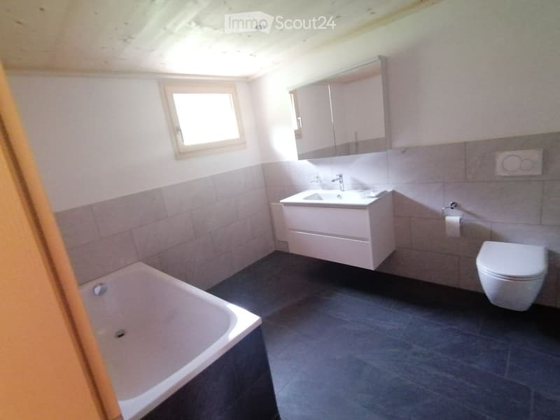 Modern bathroom with a bathtub, toilet, sink, and mirror. The walls are tiled in a two-tone design, and the floor is dark grey.