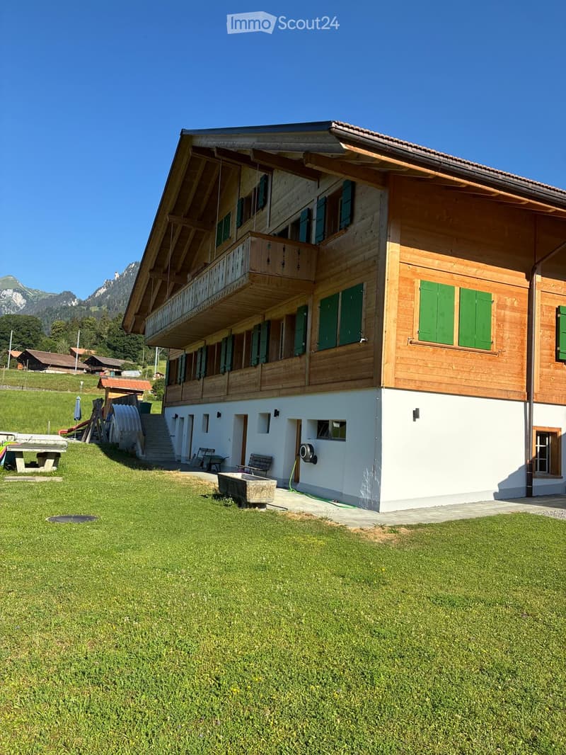 A two-story wooden house with green shutters, a balcony, and a well-maintained lawn in front. The house features a staircase leading to the entrance and is surrounded by a scenic mountain backdrop.