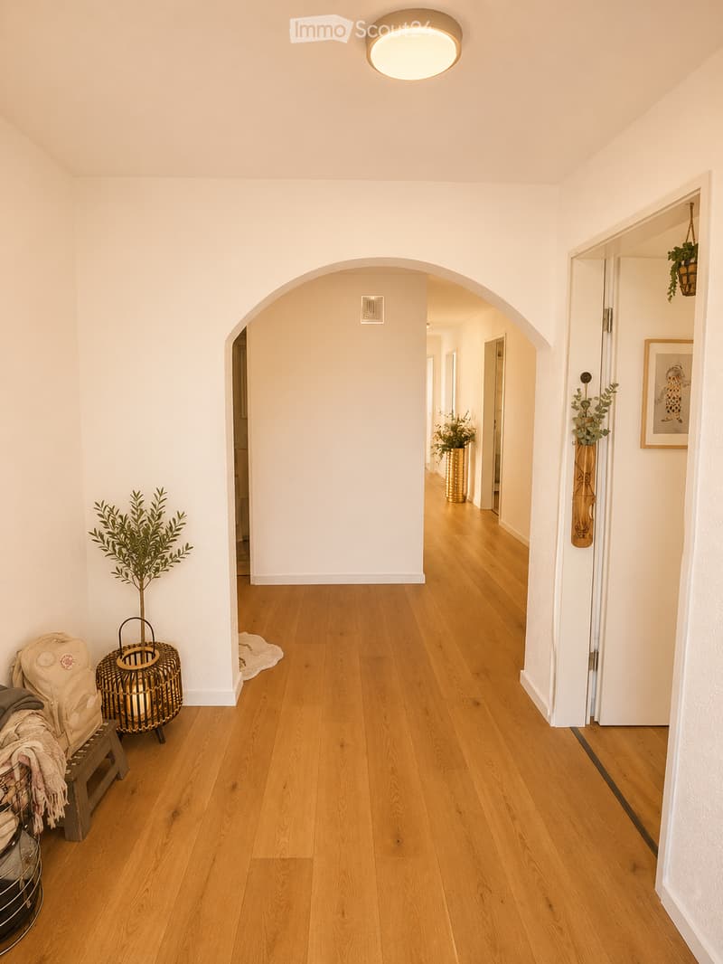 Bright hallway with wooden flooring, white walls, arched doorways, and decorative elements including a woven basket, a small bench with a blanket, and potted plants.