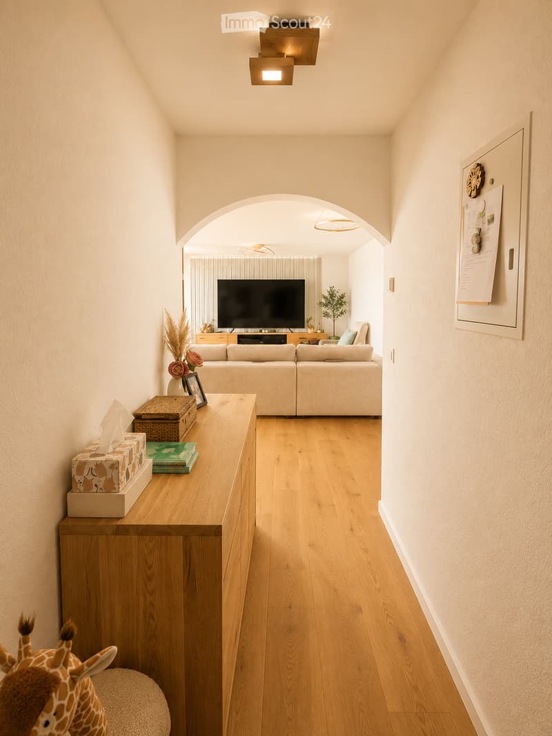 Modern living room with light wooden flooring, a sectional sofa, a television, a wooden side table with books and a tissue box, and a small giraffe toy on a round stool.