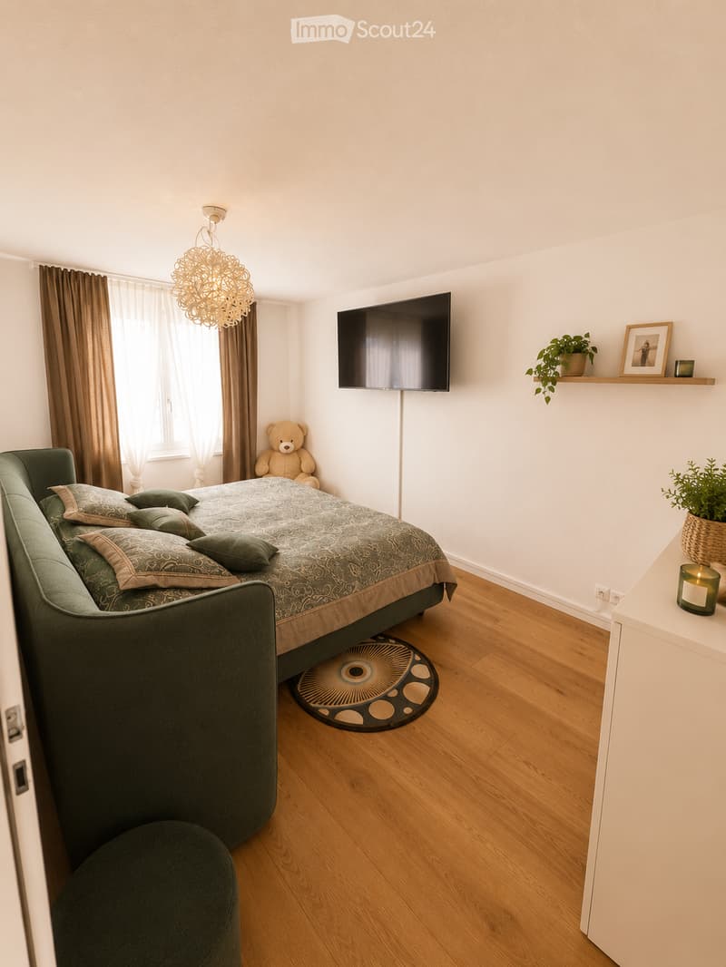 Modern bedroom with a large bed, green pillows, a round chair, a small table with a plant, a television mounted on the wall, and a decorative chandelier.