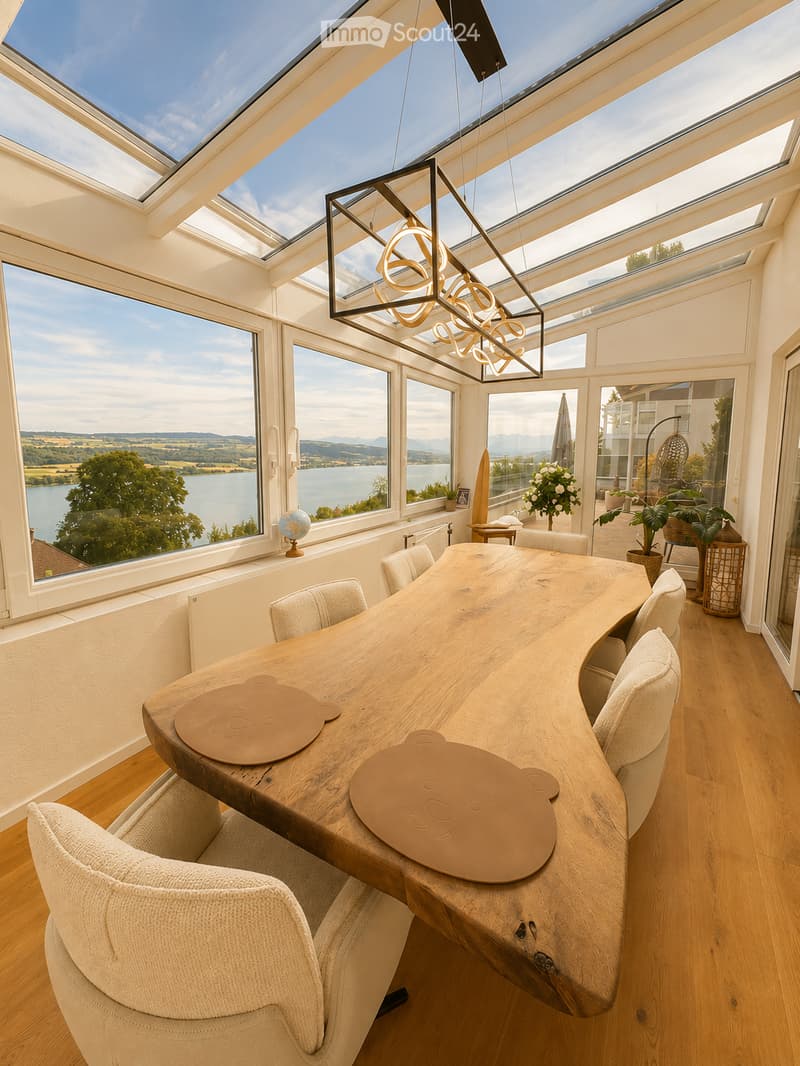 Bright, airy room with large windows and a panoramic view of a lake and surrounding landscape. Features a modern wooden table with chairs, a pendant light fixture, a globe, and potted plants.