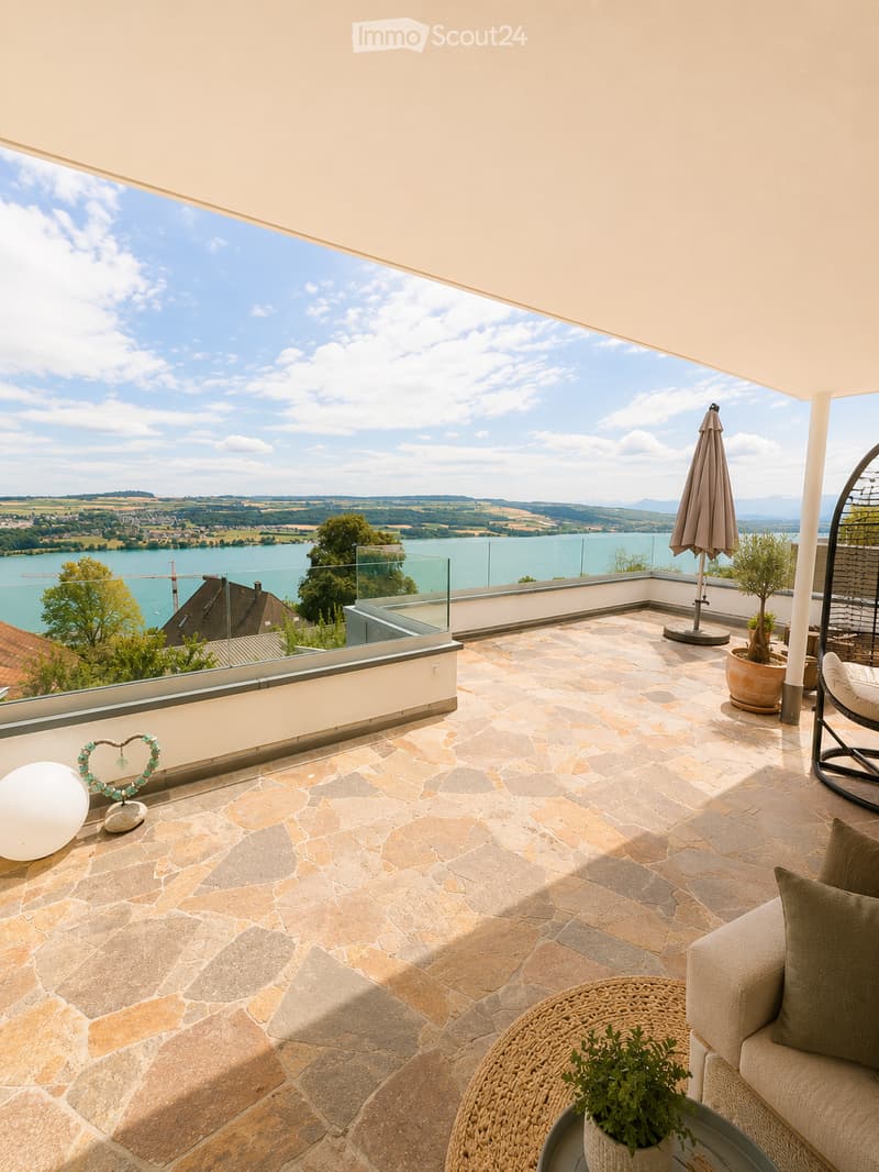 Balcony with stone tiles, glass railing, scenic lake view, sofa, table, umbrella, potted plants, and decorative items.