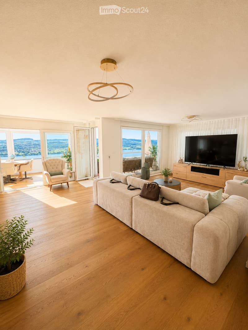 Modern living room with light wooden flooring, large windows offering scenic views, a beige sectional sofa, a coffee table, an armchair, a TV, and a dining area with chairs and a table.