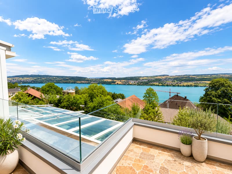 Modern rooftop terrace with glass railing, tiled floor, potted plants, and scenic lake view.