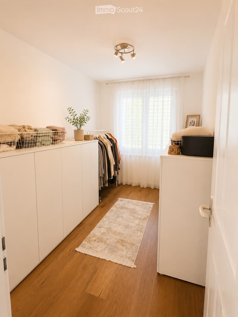 A well-lit hallway with wooden flooring, white cabinets, a hanging plant, a small rug, and neatly arranged clothing on a rack.