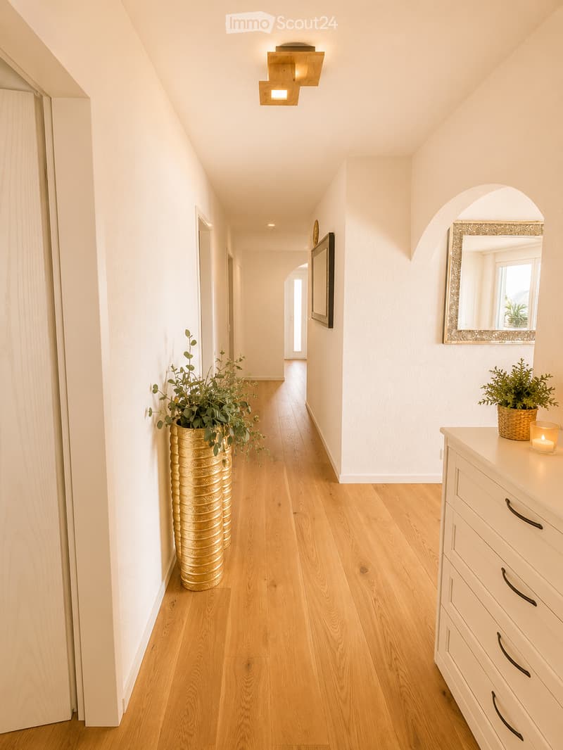 A modern hallway with light wooden flooring, white walls, and recessed lighting. Features include a large mirror, a small table with a candle, and decorative vases with greenery.