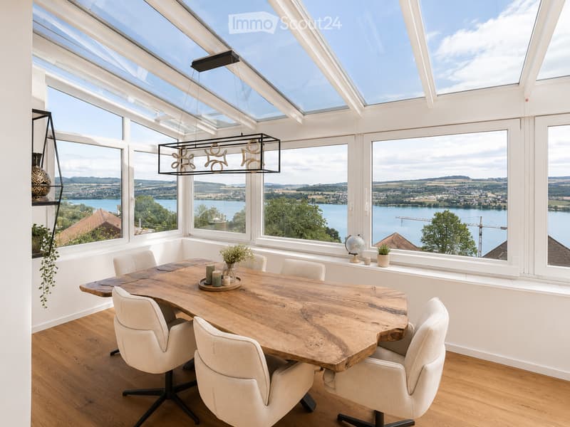 Bright dining room with large windows offering a scenic view of a lake and surrounding landscape. Features a wooden dining table with six chairs, a modern pendant light, and decorative items including a vase, candles, a globe, and potted plants.