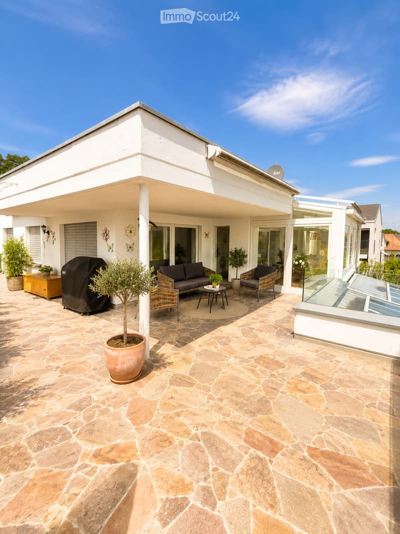 Outdoor patio with stone tiles, a sofa, a coffee table, chairs, potted plants, and a covered grill area.