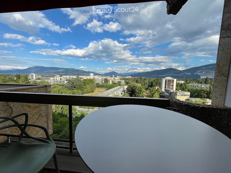 Balcony with a round table and a single chair, overlooking a cityscape with buildings and distant mountains.