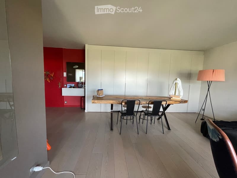 Modern, minimalist room with light wooden flooring, white cabinetry, a wooden table with chairs, a floor lamp, and a mirror in the adjacent red-walled area.