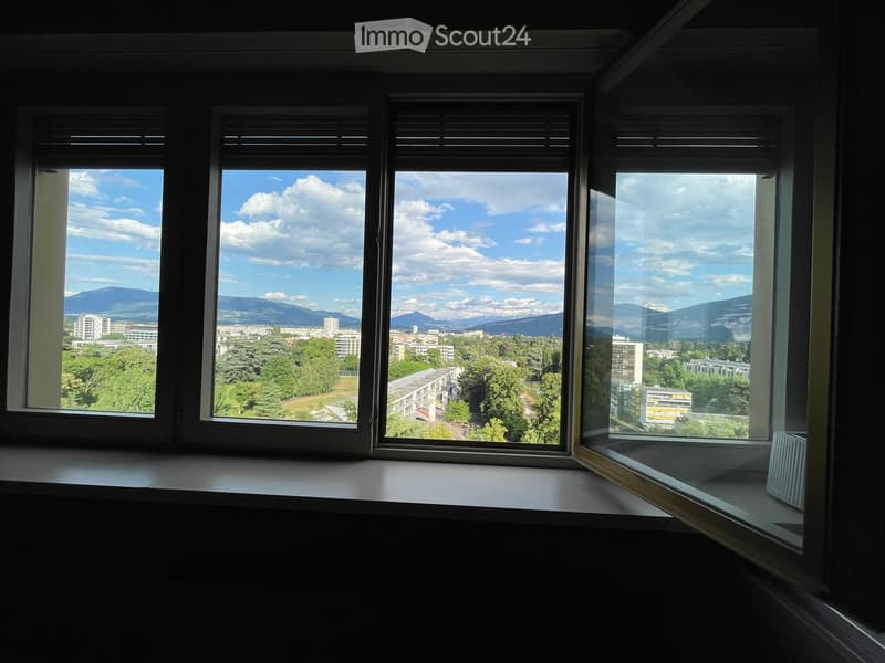 Empty room with large windows offering a scenic view of a cityscape, trees, and distant mountains.