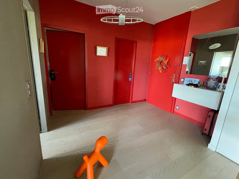 Bright hallway with red walls, light wooden flooring, and a modern white pendant light. Features a small sink area with a mirror, a decorative wreath, and an orange sculpture on the floor.