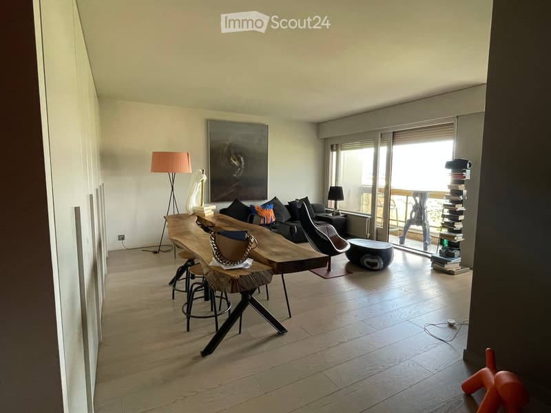 Modern living room with wooden flooring, large windows, a sofa, a wooden table, chairs, a floor lamp, a sculpture, and a bookshelf.