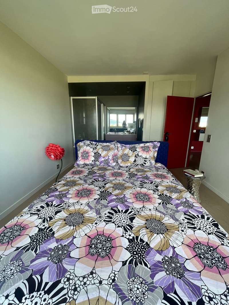 Bedroom with a large bed covered in a floral-patterned duvet, a nightstand with books, and a decorative flower on the wall.