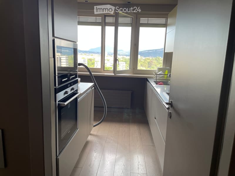 Modern kitchen with light wooden flooring, white cabinets, stainless steel oven, and a sink with a faucet. Large windows offer a scenic view of the city and mountains.