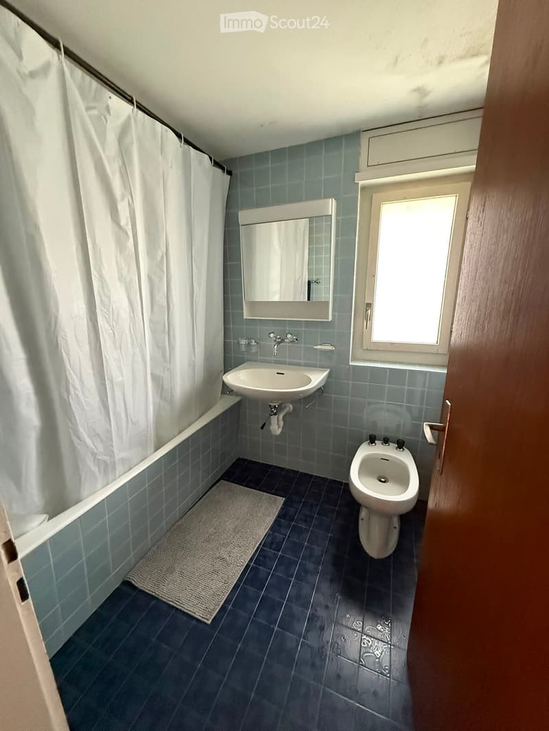 Bathroom with blue tiled walls, bathtub, sink, bidet, mirror, and a small window. Shower curtain is visible on the left side.