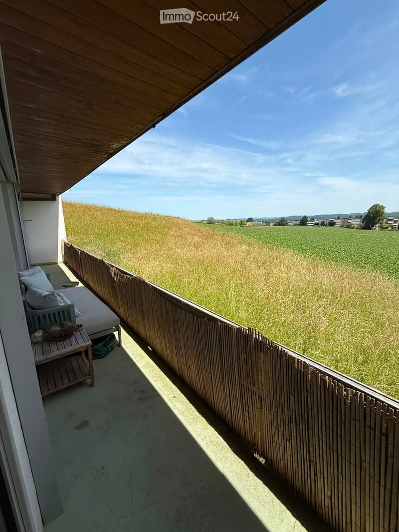 Balcony with wooden railing, sofa, small table, and view of a green field.