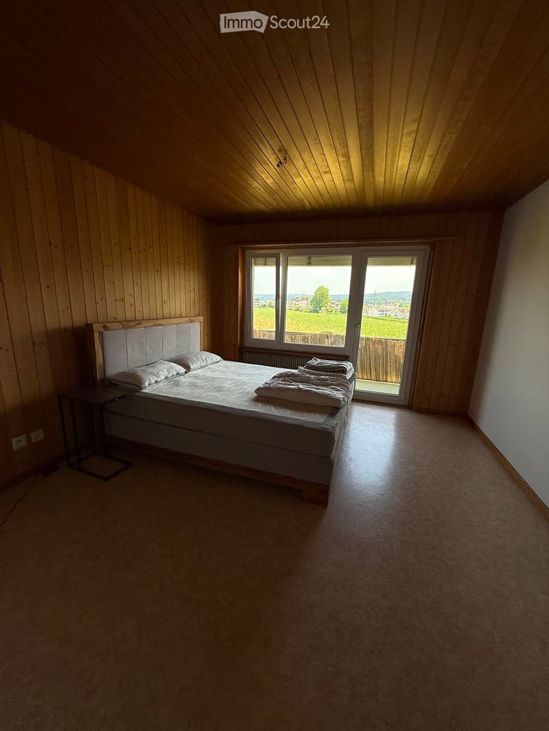 Cozy bedroom with wooden walls and slanted ceiling, featuring a double bed with pillows and blankets, a nightstand, and a large window offering a view of the countryside.