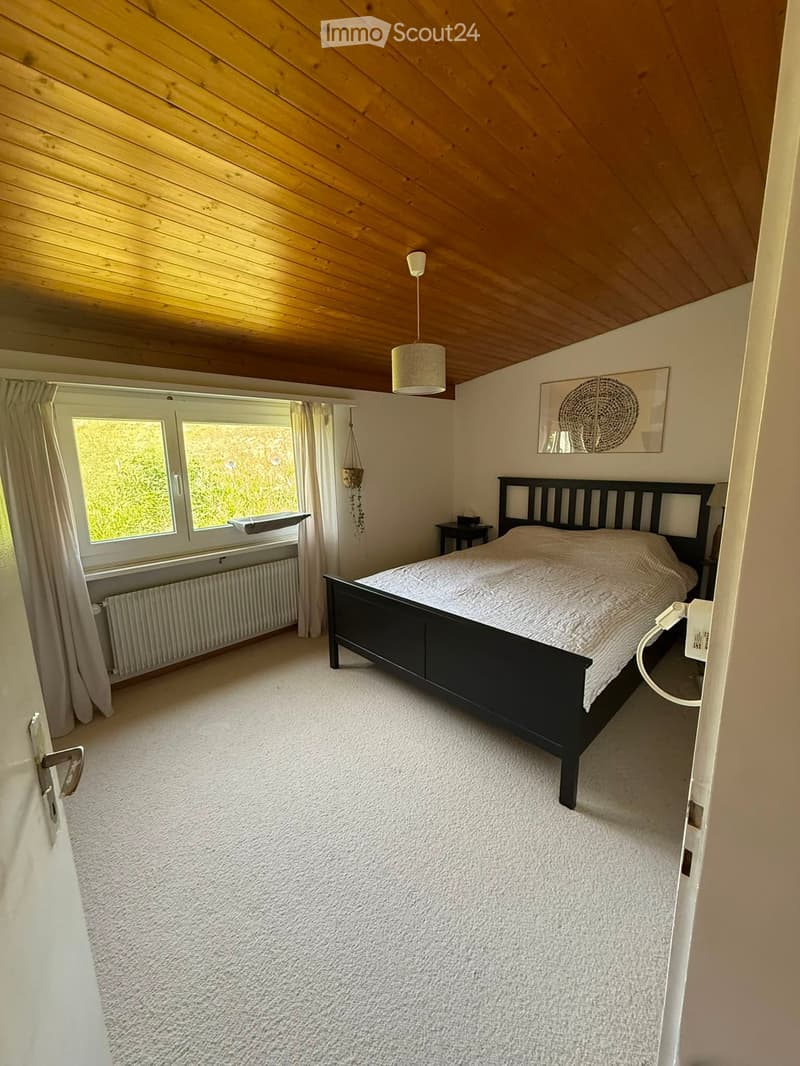 Bright bedroom with a wooden slanted ceiling, white walls, large windows with curtains, a double bed with a dark wooden frame, a bedside table, and a hanging plant.