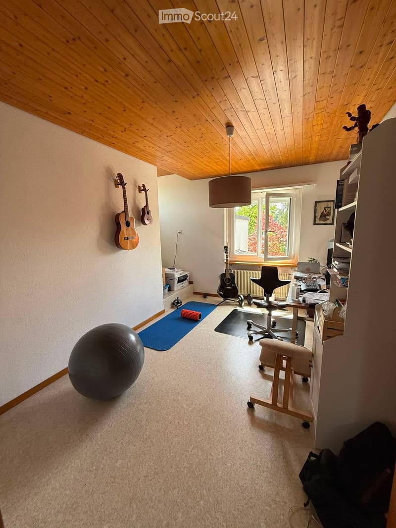 A multi-purpose room with a slanted wooden ceiling, large window, and various items including guitars, a chair, a ball, a table with a laptop, and a stool.