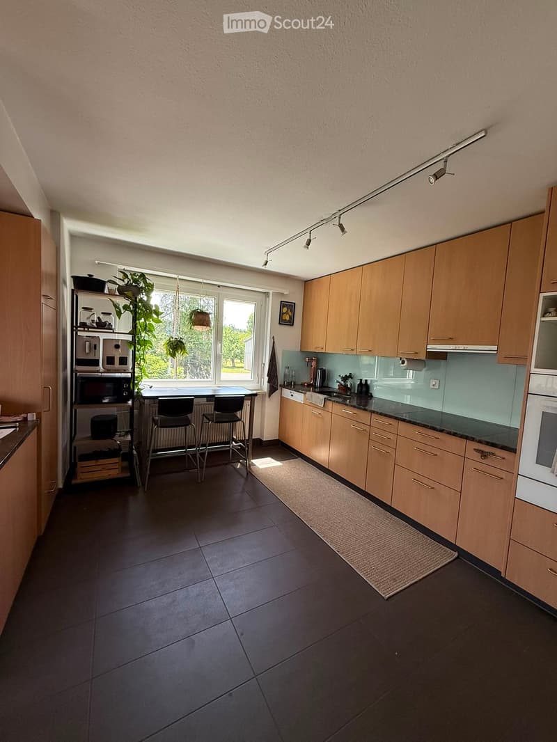 Modern kitchen with light wood cabinetry, dark tiled floor, island with stools, and large windows overlooking a green area.
