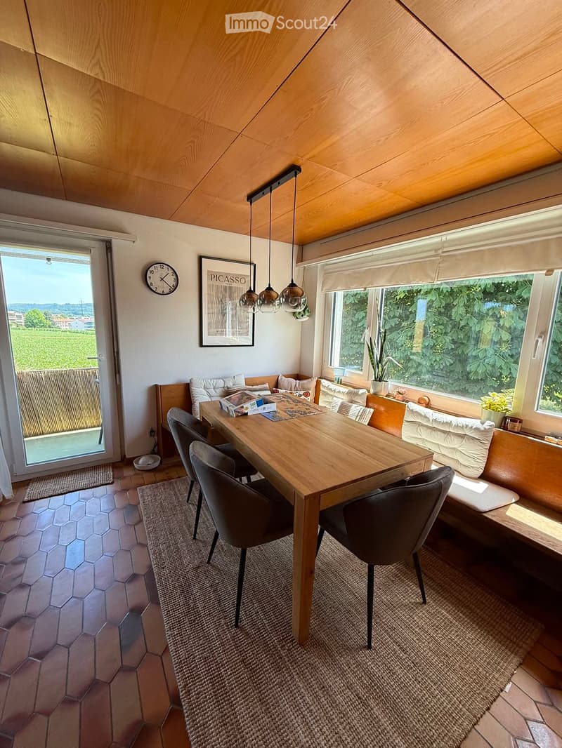 Bright, modern dining area with wooden table, chairs, built-in sofa, pendant lights, and large windows overlooking a green landscape.