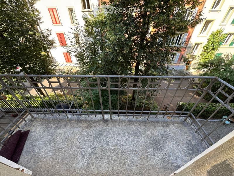 Balcony with metal railing, concrete floor, and view of neighboring buildings and greenery.