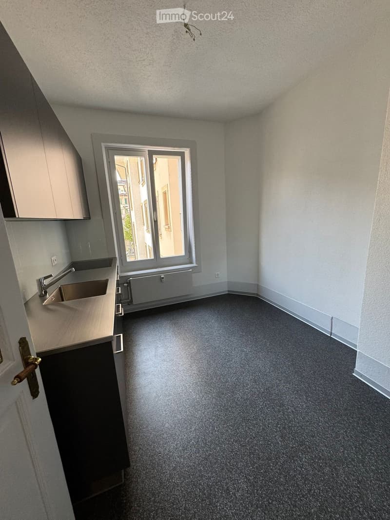 Empty room with grey carpet, white walls, a small kitchen area with sink and cabinets, and a large window with blinds.