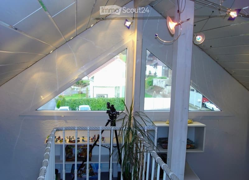 A well-lit room with large windows, a small table with toys, shelves, a potted plant, and a camera on a tripod. The room features modern lighting fixtures and a white railing.