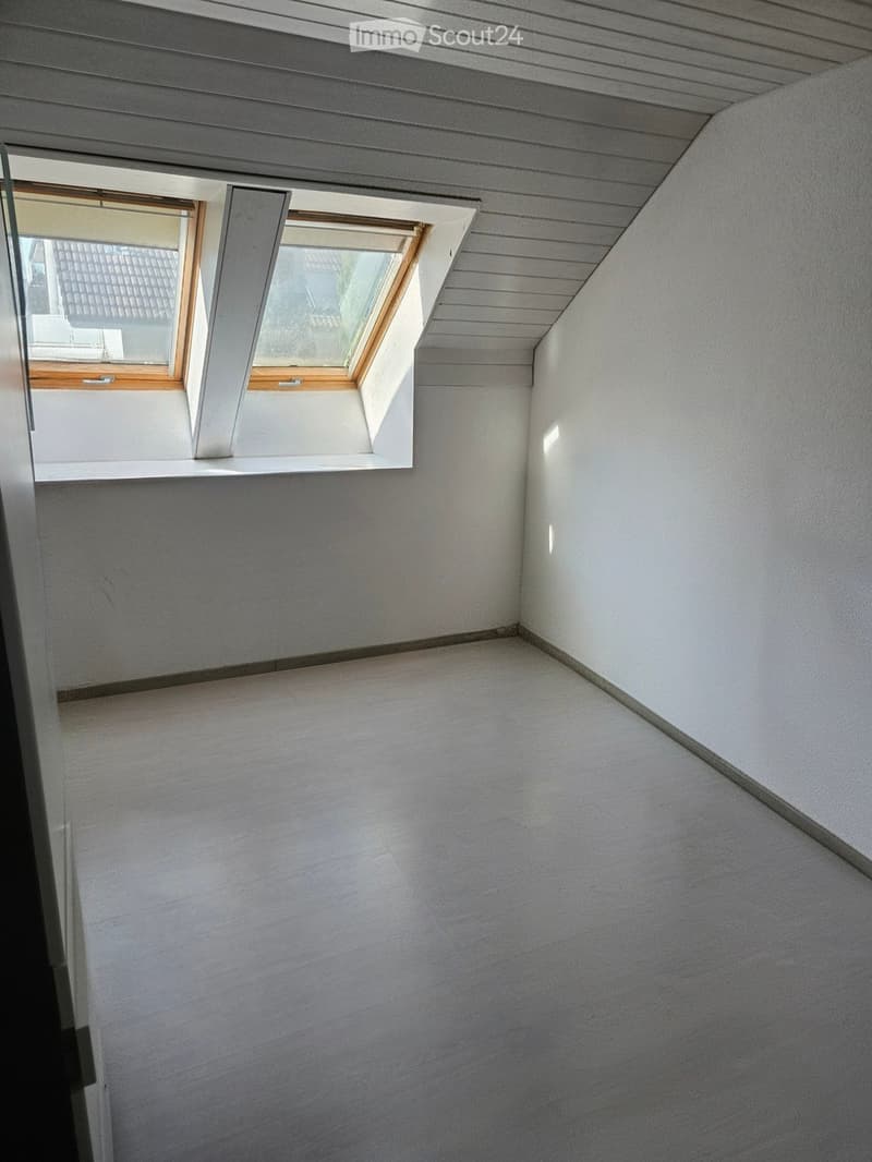 Empty room with light grey walls and a wooden floor, featuring a skylight window and a grey ceiling with wooden paneling.