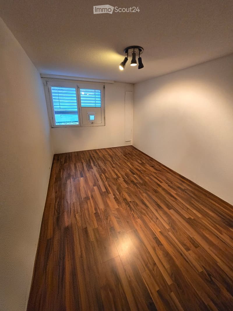 Empty room with wooden flooring, white walls, and a small window with blinds. A ceiling light fixture is visible.