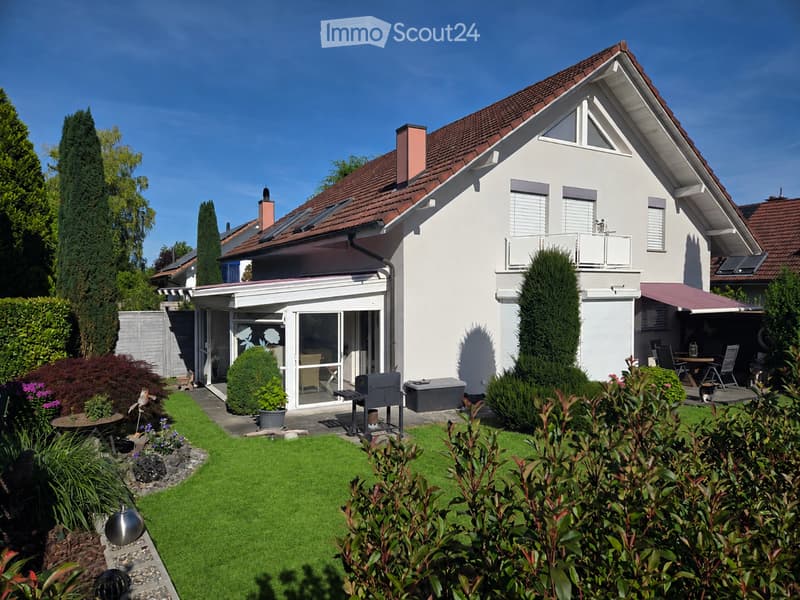 Two-story house with white exterior, red-tiled roof, and a well-maintained garden featuring a lawn, shrubs, and a patio area with outdoor furniture.