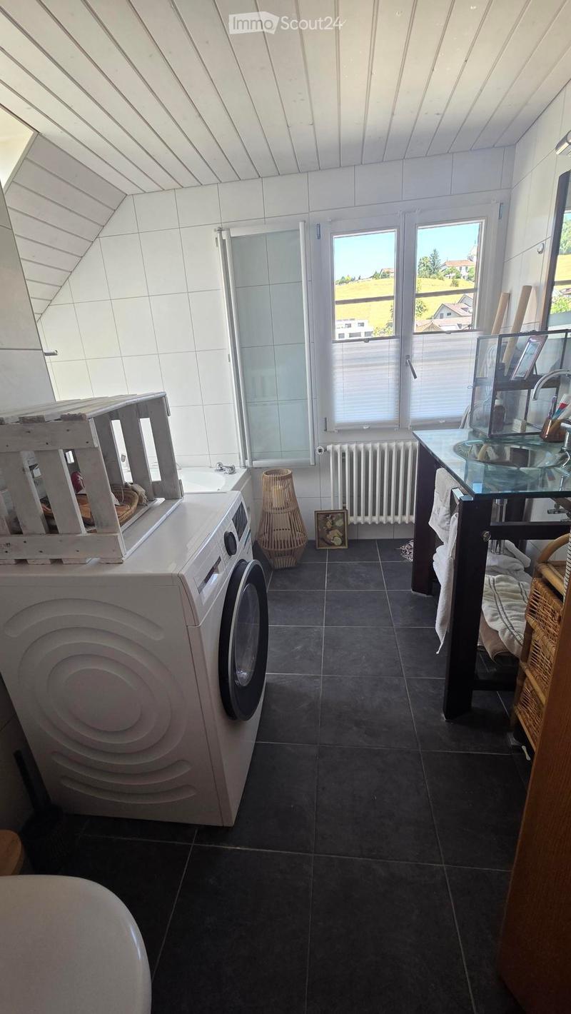 Bathroom with a washing machine, sink, mirror, radiator, wicker basket, and a picture frame. The room has dark tiled flooring and white tiled walls, with a window offering a view of the outdoors.