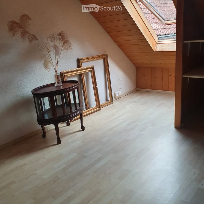 Attic room with wooden floors, slanted wooden ceiling, and skylight. Features a small round table with a vase and two large picture frames leaning against the wall.