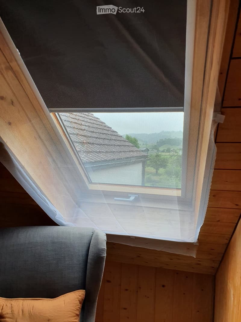 A cozy living room with wooden walls and a large window with a view of the countryside. A grey sofa with a cushion is visible in the foreground.