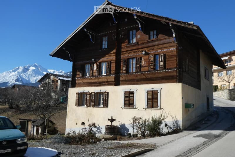 Traditional wooden house with a raised ground floor, multiple windows, and a view of mountains in the background.