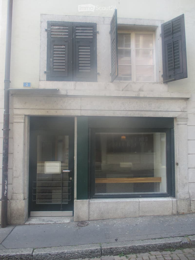 Facade of a building with a glass door entrance, a window with shutters, and a small plaque above the door.