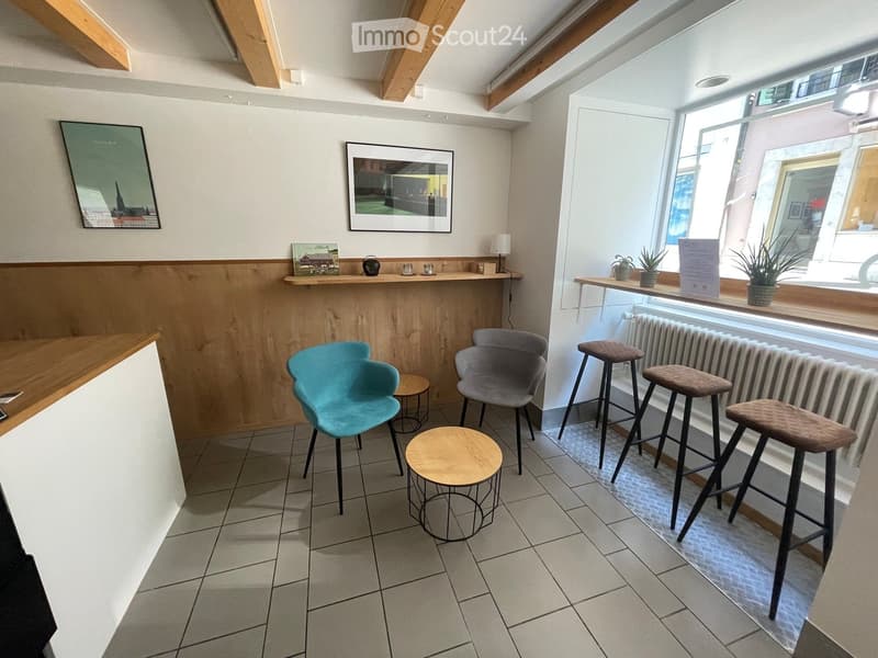 A modern, well-lit room with exposed wooden beams, tiled flooring, and a mix of seating options including stools and chairs. There are small tables, a lamp, and decorative items on shelves and windowsills.