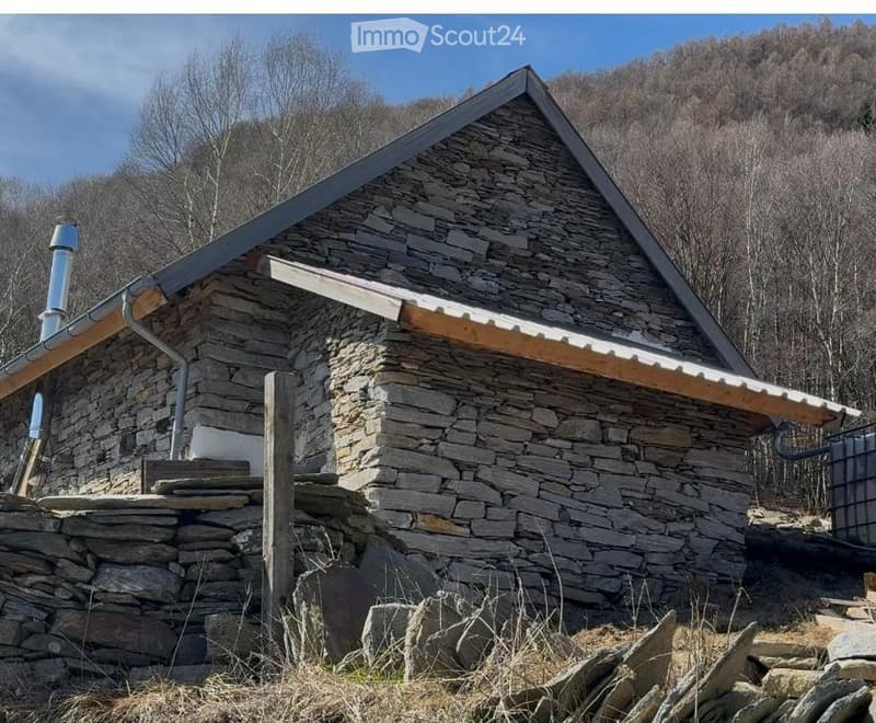 Stone cottage with a pitched roof, wooden trim, and chimney, situated in a rural, forested area.