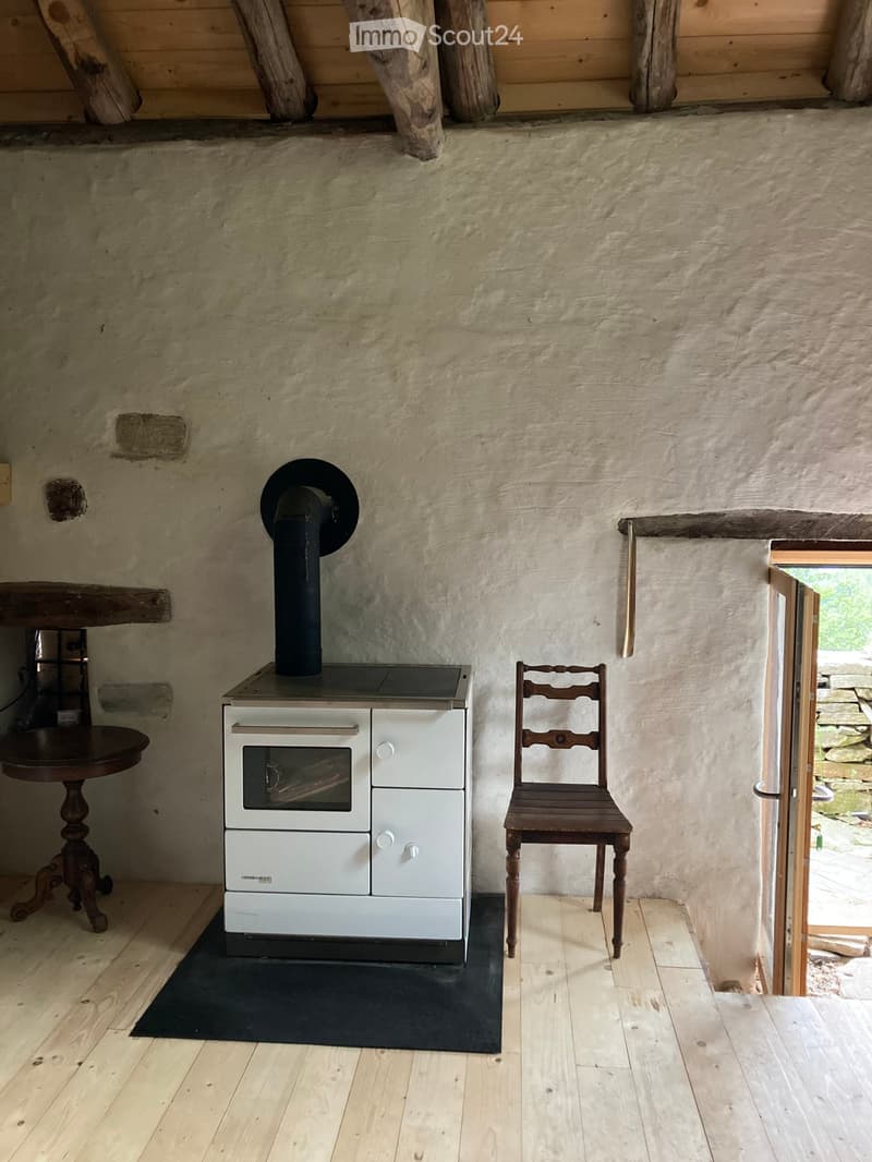 A rustic room with a wood stove, a wooden chair, and a small table, featuring exposed wooden beams and a stone wall.