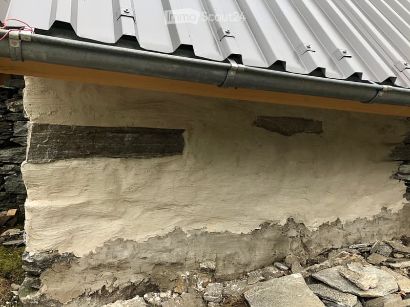 Close-up view of a weathered building facade with a stone base, plaster wall, and metal roof overhang. The wall shows signs of wear and discoloration, with visible cracks and patches.