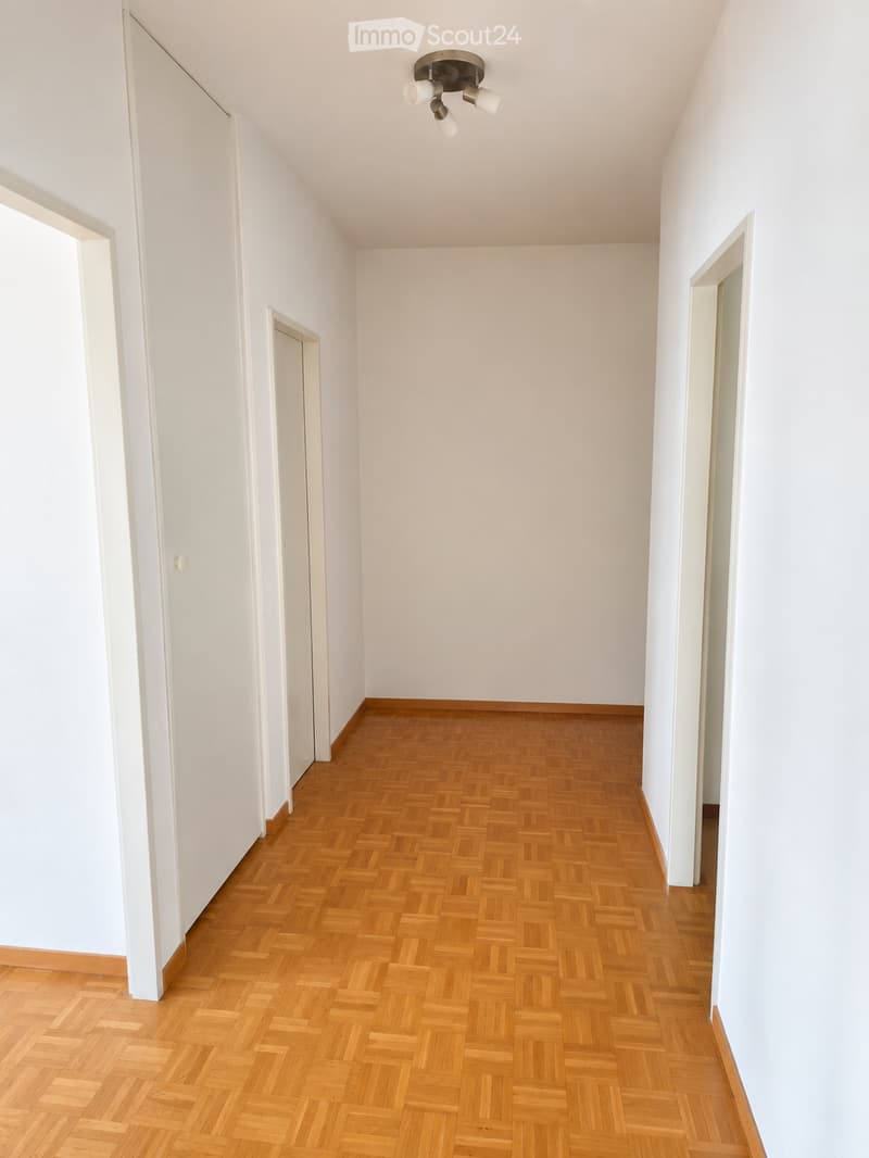 Empty hallway with wooden herringbone flooring and white walls. Features a ceiling light fixture and several doorways.