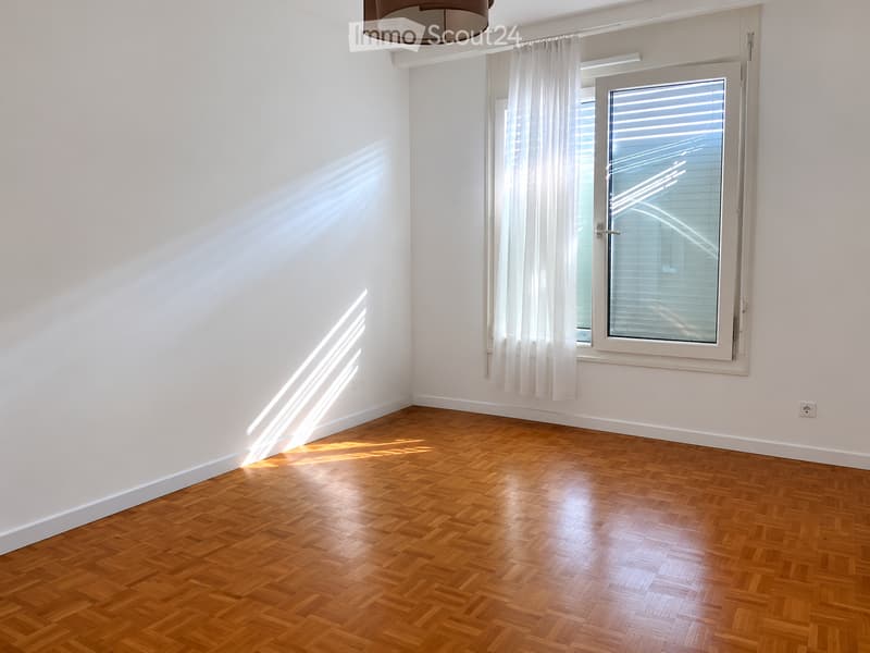 Empty room with wooden herringbone flooring, white walls, and a large window with white curtains. Sunlight streams in, casting shadows on the floor.
