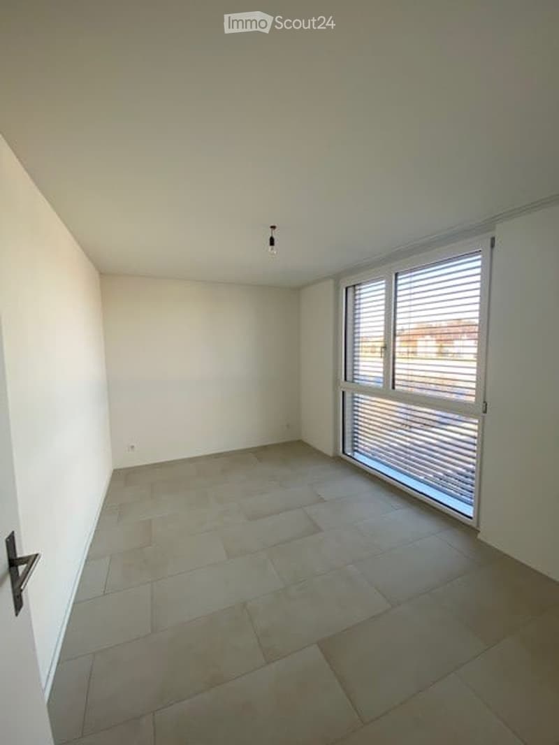 Empty room with light-colored tiled flooring, white walls, and a large window with blinds. The room has a single door and a ceiling light fixture.