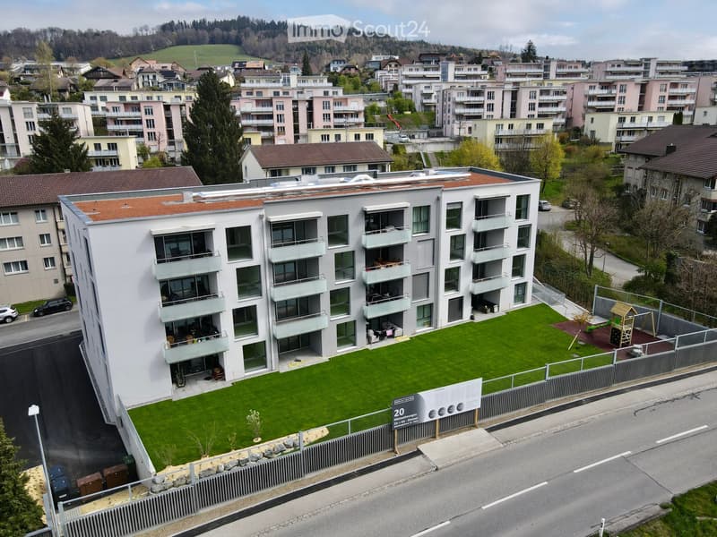 Modern multi-story residential building with balconies, surrounded by a green lawn and playground area. The building features large windows and a contemporary design.