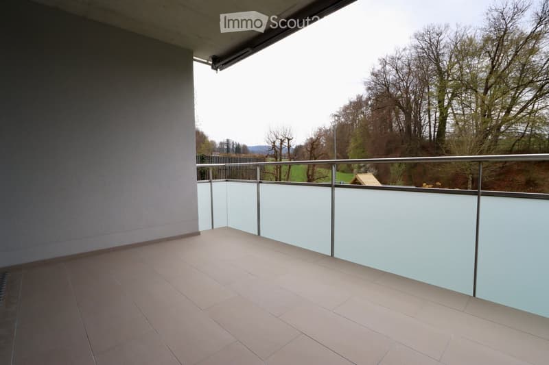 Balcony with tiled floor, glass railing, and a view of trees and a small structure in the distance.