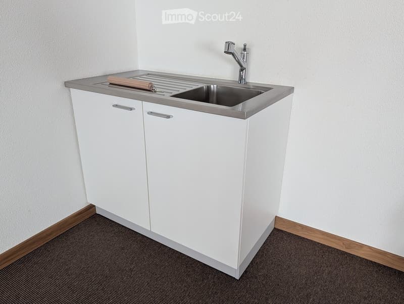 Modern white kitchen cabinet with a stainless steel sink and faucet, placed against a white wall with a dark carpeted floor.